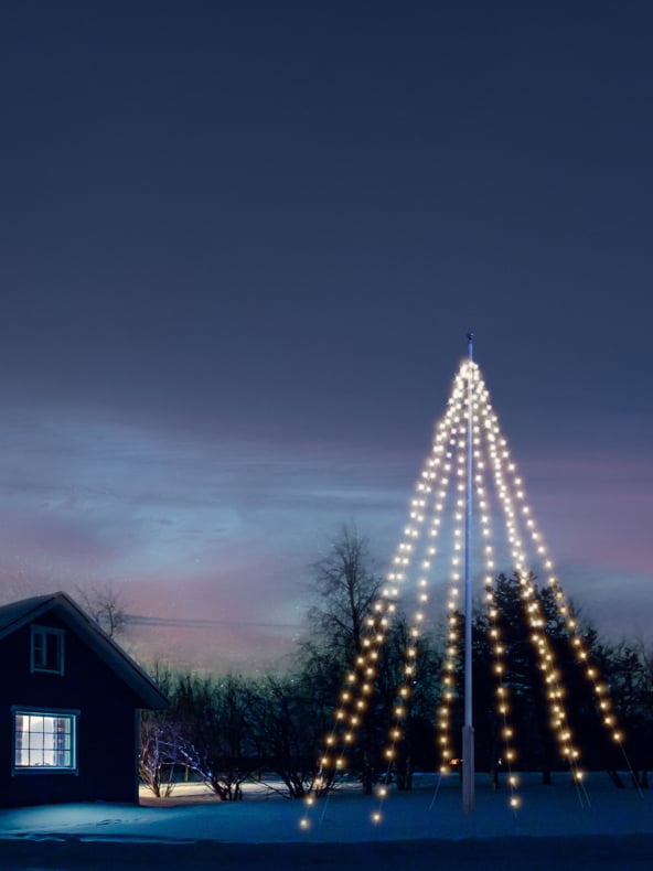 Reihen weißer Lichter bilden eine Baumform auf einem hohen Pfahl draußen; Schneebedeckten Boden und ein Haus mit einem leuchtenden Fenster bei Einbruch der Dämmerung.