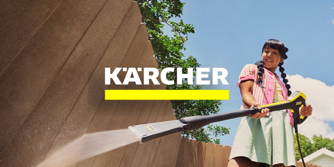 Kobieta czyści drewniane ogrodzenie na zewnątrz myjką ciśnieniową Kärcher; logo Kärcher i żółta linia widnieją na środku.