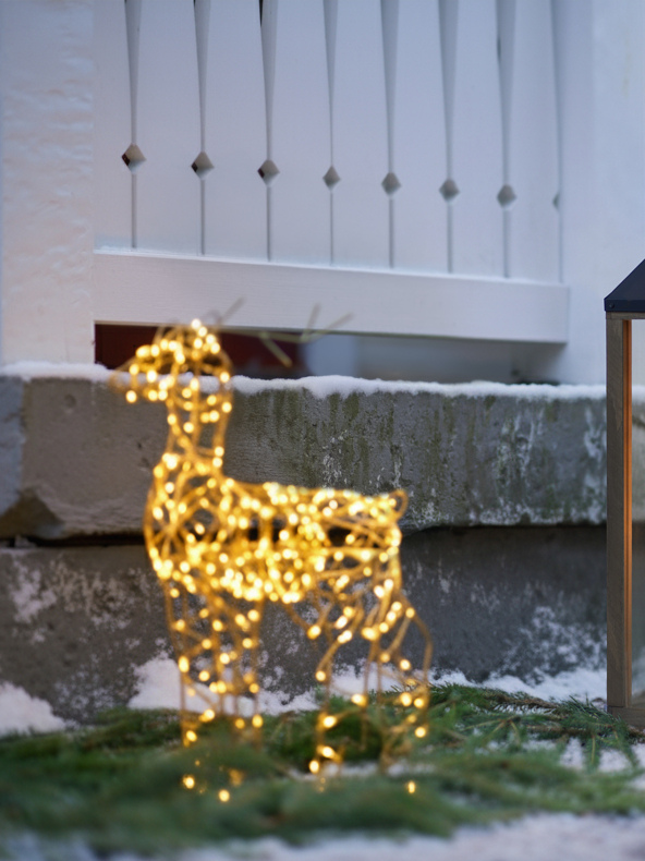 Eine mit gelben Lichtern bedeckte Rentierdrahtskulptur steht auf schneebedecktem Grün neben einem weißen Zaun und grauen Steinstufen.