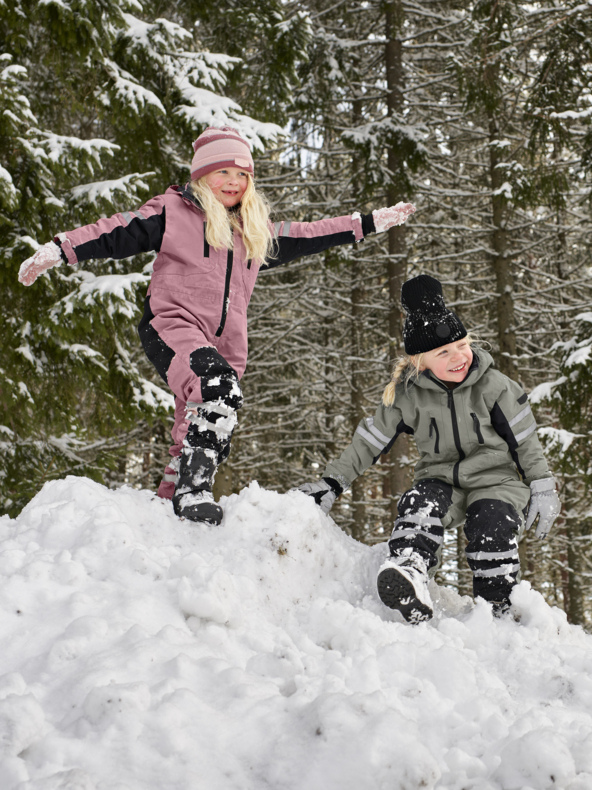 Barn i vinterkläder leker på en snötäckt kulle bland träd; Den ena balanserar med armarna utsträckta, den andra hukar sig tätt intill.