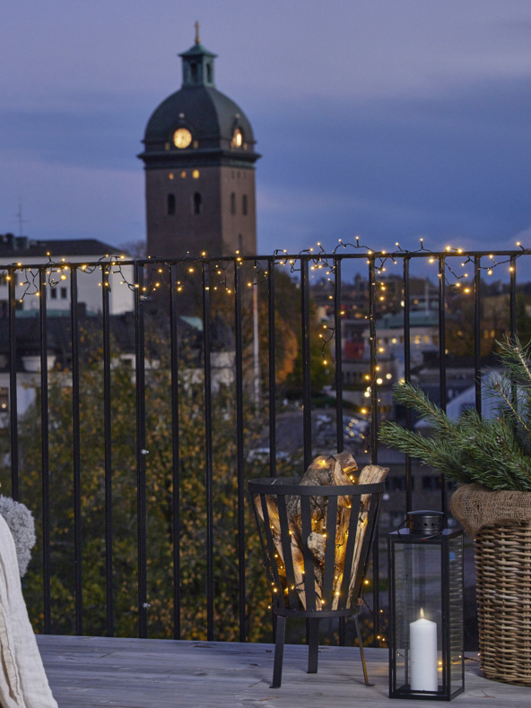 Dachterrasse mit Lichterketten, Brennholzhalter, Glaslaterne bei Kerzenschein, grünen Zweigen und Skyline der Stadt mit Uhrturm in der Abenddämmerung.
