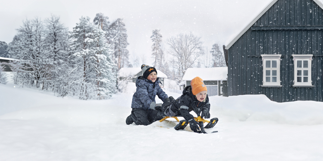 Två barn i vinterkläder leker i snön, den ena skjuter en släde och den andra åker. Ett mörkt hus och snötäckta träd ligger bakom dem.