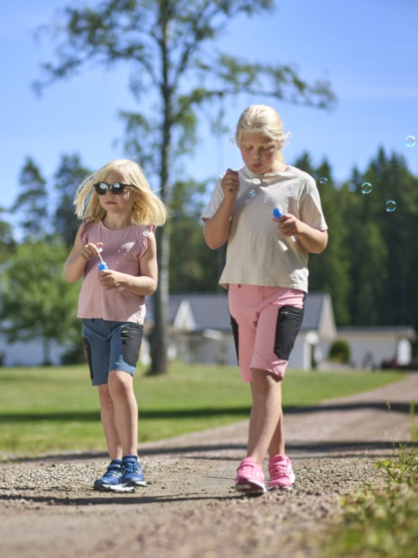 Två barn går på en grusväg och blåser bubblor. Man bär solglasögon, rosa skjorta, blå shorts; Den andra en ljus topp och rosa shorts. Träd och hus finns bakom dem.