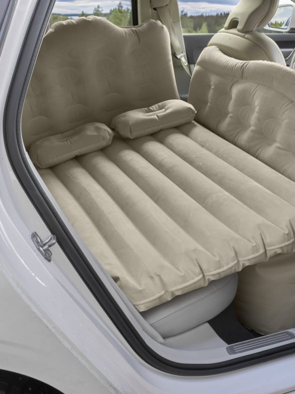 Eine beige aufblasbare Luftmatratze mit zwei Kissen bedeckt den Rücksitz eines weißen Autos und bildet einen flachen Raum über der Beinfreiheit.