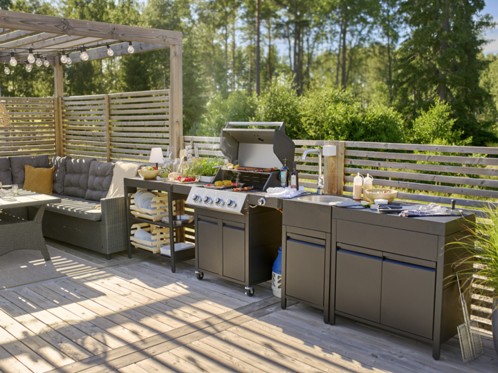 En träterrass med en gasgrill, bänkskiva, förvaringsskåp, utomhussittplatser, avskärmningsskärmar och träd bortom.