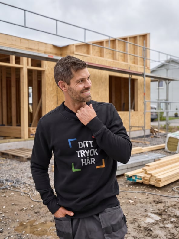 En man ler och tittar åt sidan utanför ett trähus som byggs, iklädd en svart sweatshirt med "DITT TRYCK HÄR" och färgade konsoler.