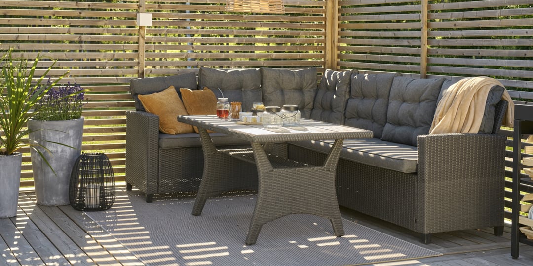 Rottet terrassesofa med grå puter, to gule puter og bord dekket med drikke. Planter, lykt og trelist som bakgrunn.
