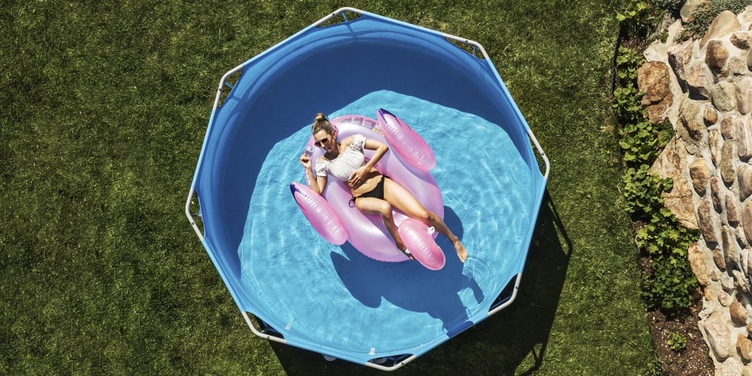 En kvinna slappnar av på en rosa flotte i en liten blå pool placerad på gräs bredvid en stenmur, sedd från ett översiktsperspektiv.