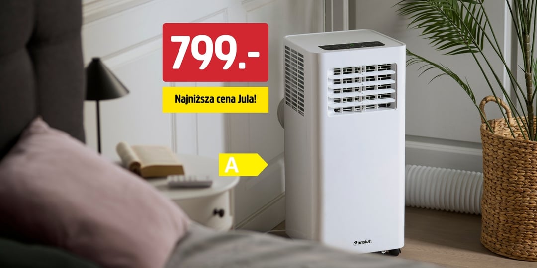 Na podłodze w salonie, obok rośliny i sofy, stoi przenośny biały klimatyzator. Na tabliczce widnieje napis „799,- Najniższa cena Jula!” wraz z żółtym symbolem litery „A”.