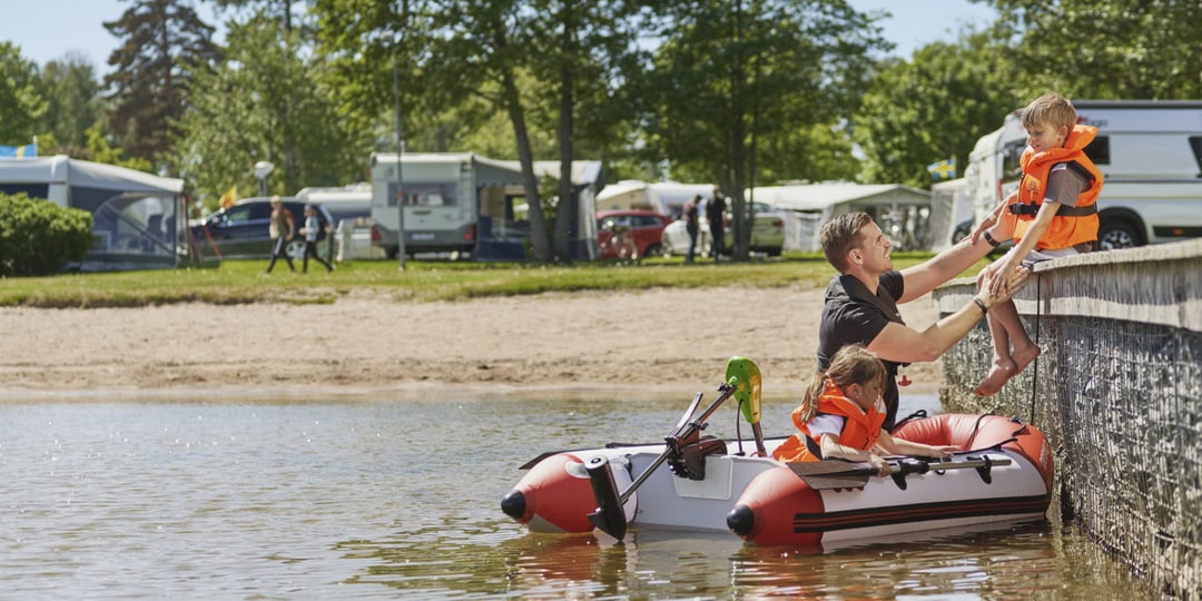 Ein Erwachsener hebt ein Kind in einer orangefarbenen Schwimmweste von einem Floß auf einen Steg, während ein anderes Kind wartet. Wohnmobile, Bäume und Menschen liegen hinterher.