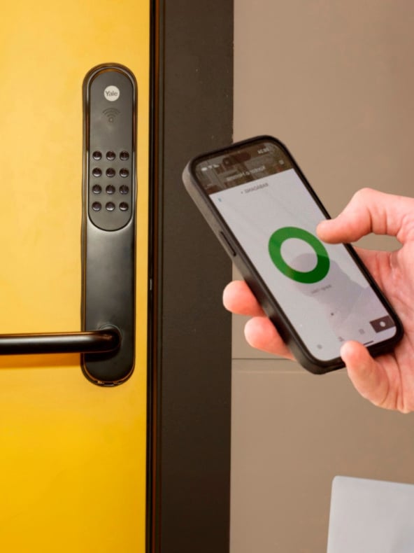 Noen bruker en smarttelefon som viser en grønn sirkel ved siden av et svart tastaturlås på en gul dør, noe som indikerer at telefonen låser opp døren elektronisk.