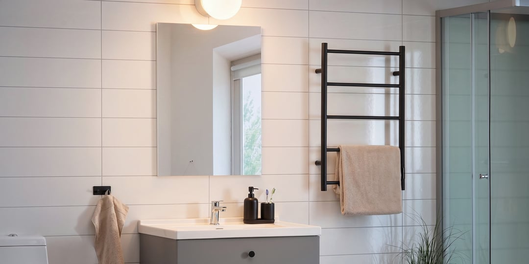 Stilrent badrum med en rektangulär spegel över handfatet, ett svart handduksställ med en beige handduk, ljusgrå kaklade väggar och en glasdusch till höger.