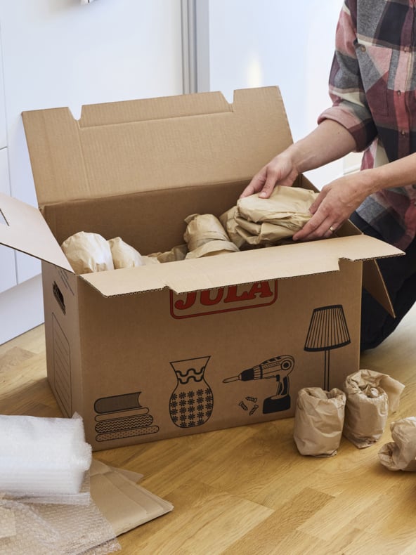 Med en rutig skjorta lägger man inslagna tillhörigheter i en stor flyttlåda av kartong på ett trägolv. Lådan visar bilder på böcker, en vas, en lampa och en borrmaskin. Ark med bubbelplast är nära till hands.