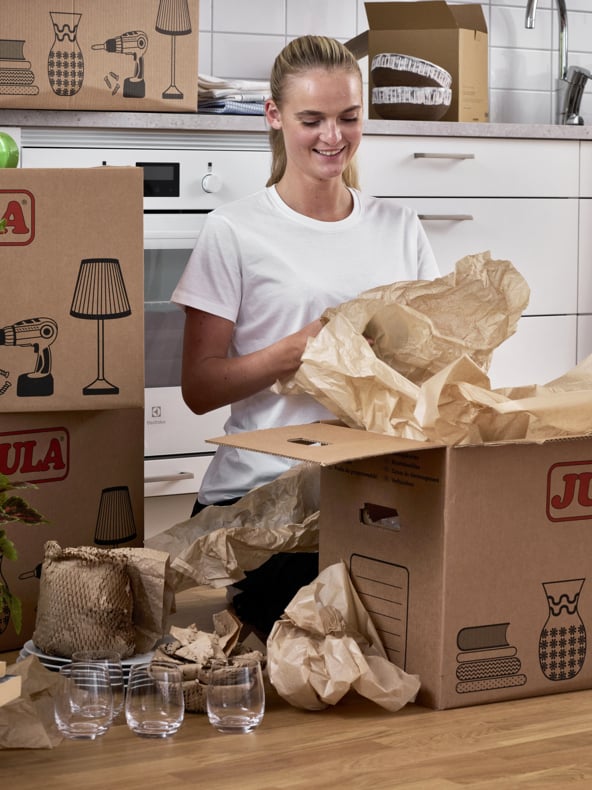 En kvinna i en vit t-shirt packar upp kartonger i ett kök. Märkta lådor, packpapper, tre klara glas och några inslagna föremål ligger utspridda på bänken och golvet.