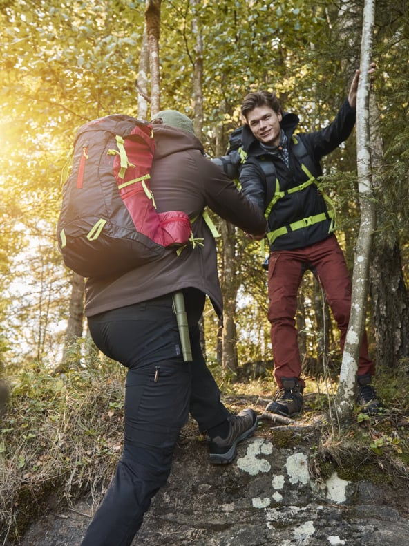 Två vandrare tar sig genom en solig skog, där den ena hjälper den andra uppför en kort stenig sluttning. De är klädda i utomhuskläder och bär ryggsäckar medan solstrålar lyser genom träden och lyser upp naturen.