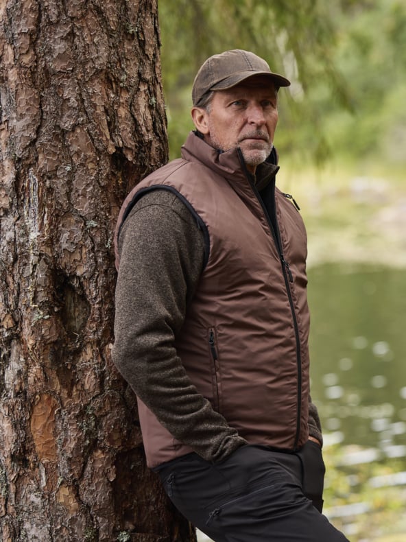 En man i brun keps, brun väst och mörka kläder lutar sig tillbaka mot ett träd, vänd mot kameran. Frodig grönska omger honom, med ett mjukt suddigt vattendrag i fjärran.