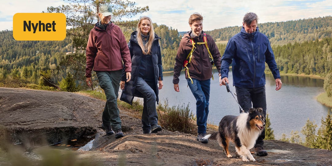 Fyra personer och en hund går längs en stenig stig vid en sjö och genom skogen, alla klädda i jackor och byxor. En person håller i hundens koppel. I det övre vänstra hörnet finns en gul skylt med texten ”Nyhet”. Himlen är delvis molnig.