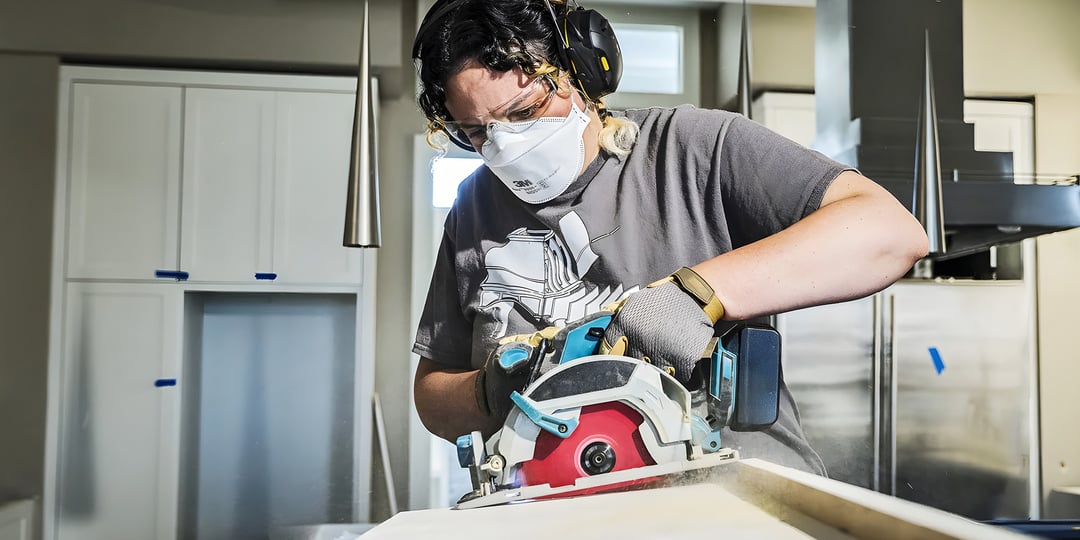 En person som bär skyddsglasögon, hörselskydd, handskar och en mask använder en cirkelsåg för att såga i trä inomhus. Bakom personen syns vita skåp och silverfärgade vitvaror.