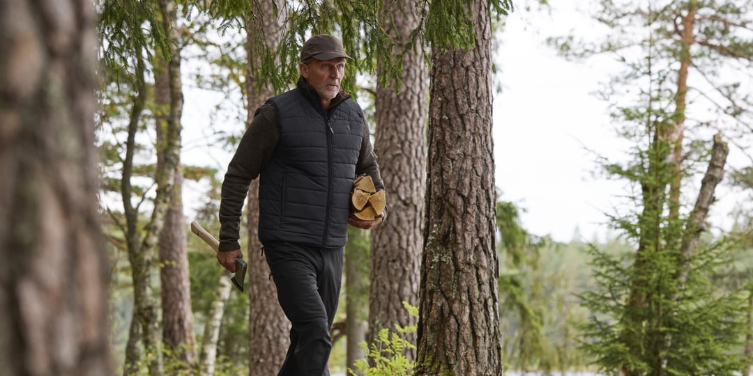 En mann iført vest og lue går gjennom en skog med en bunt ved og en øks.