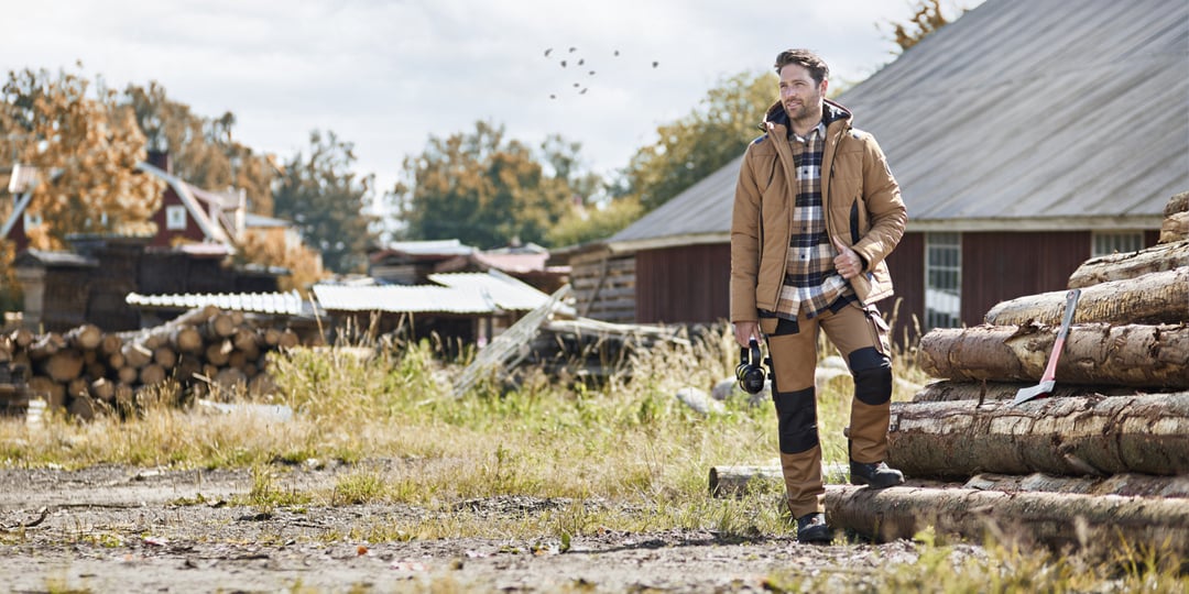 En man i en beige arbetsjacka, rutig skjorta och förstärkta byxor står bredvid en hög med stockar med en yxa vilande ovanpå.
