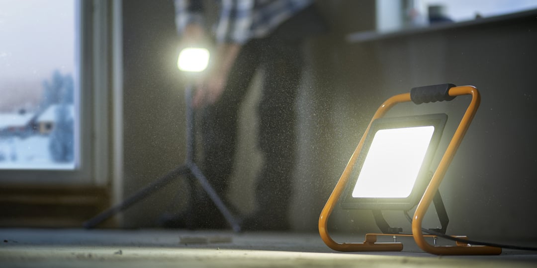 Zwei tragbare LED-Arbeitslampen beleuchten eine Baustelle, auf der im Hintergrund eine Person arbeitet.