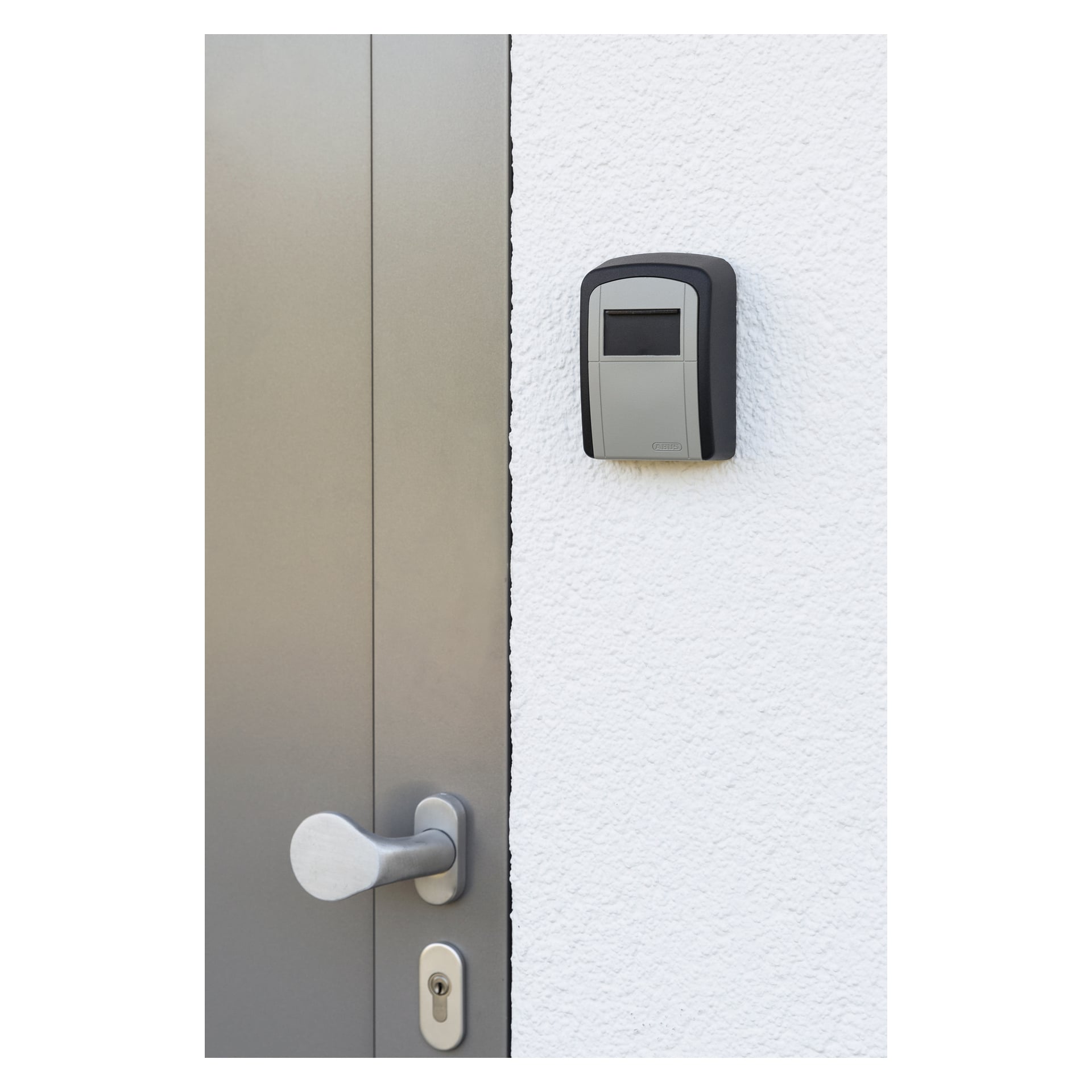 En sølvfarget dør med sylindrisk håndtak og nøkkelhull står ved siden av en vegg med hvit tekstur. Montert over håndtaket er et grått og svart ABUS Key Garage 707 nøkkelskap.