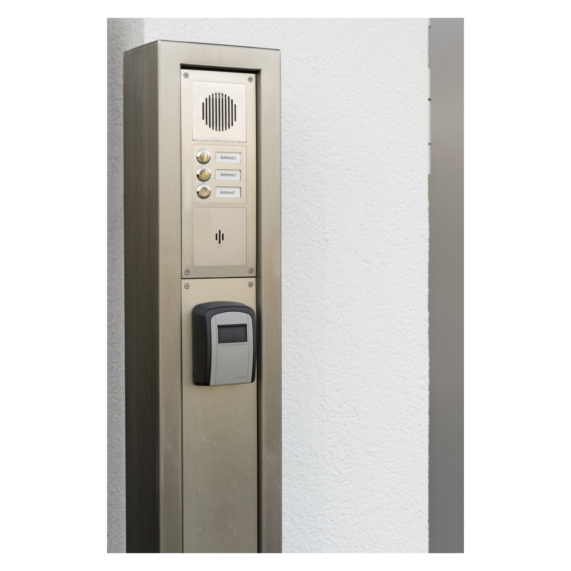 Et ABUS Key Garage 707 nøkkelkabinett er montert på en vertikal stolpe, og viser et metallpanel utstyrt med tre merkede knapper, en høyttaler og en kortleser.