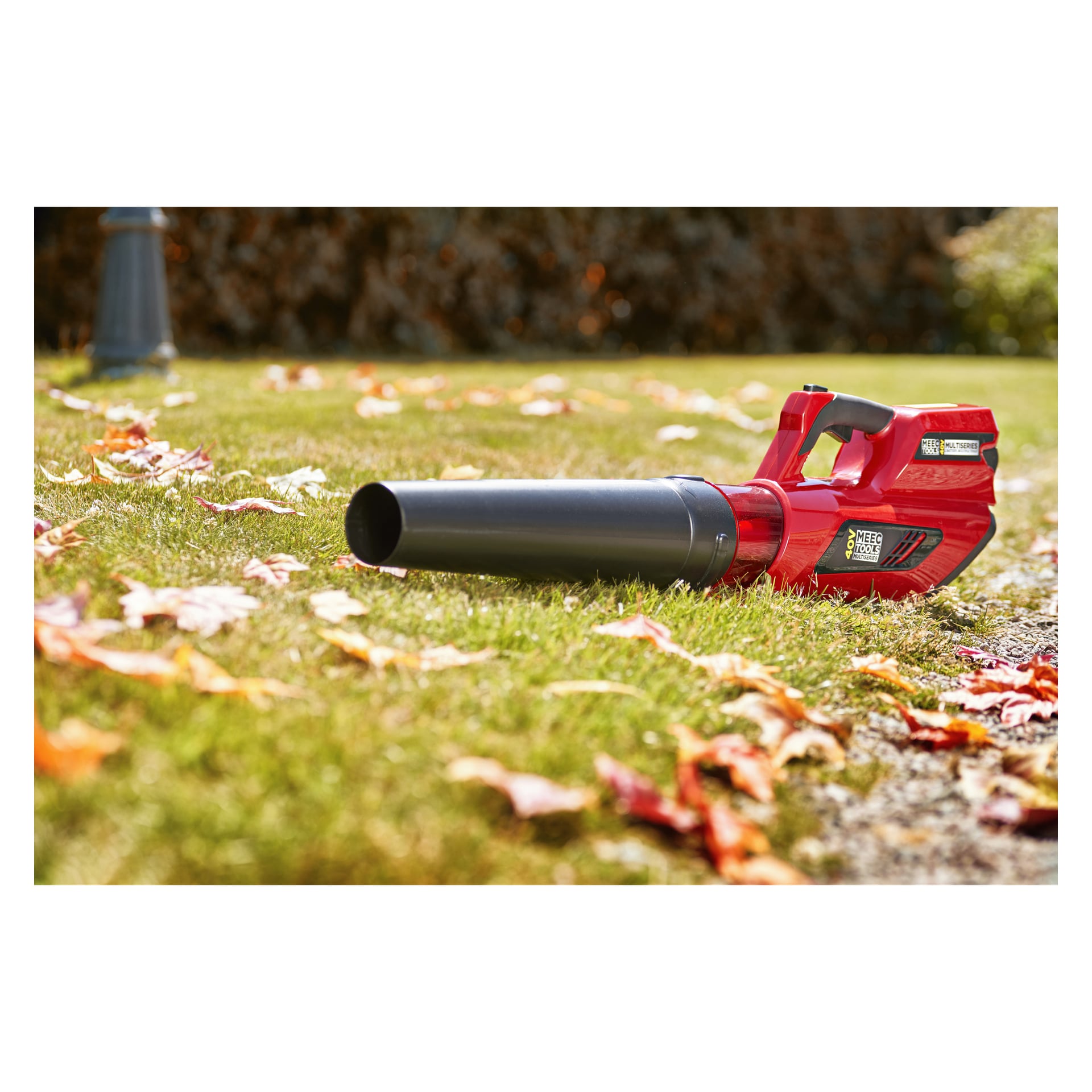 En rød og svart Meec Tools Multiseries 40 V batteridrevet løvblåser står på grønt gress blant spredte høstløv. Den mykt uskarpe bakgrunnen viser flere blader og noen busker.