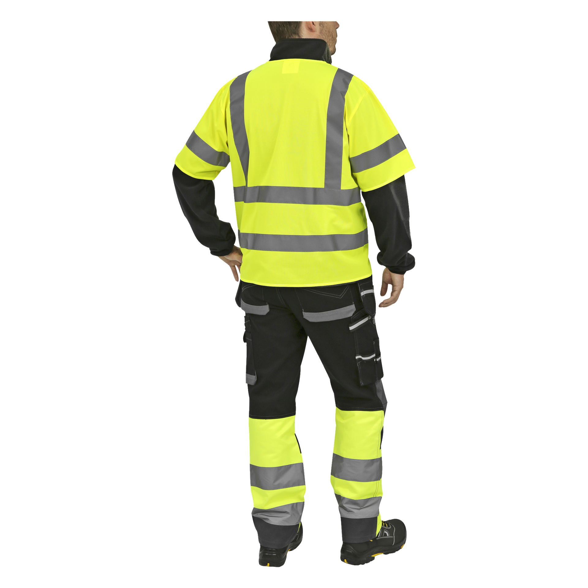 En person vänder blicken bort från kameran, klädd i en synlig gul och svart Bluewear reflekterande väst, verktygsbyxor med reflekterande detaljer och svarta skor. De har höger hand på höften och vänster arm vilande löst vid sidan.