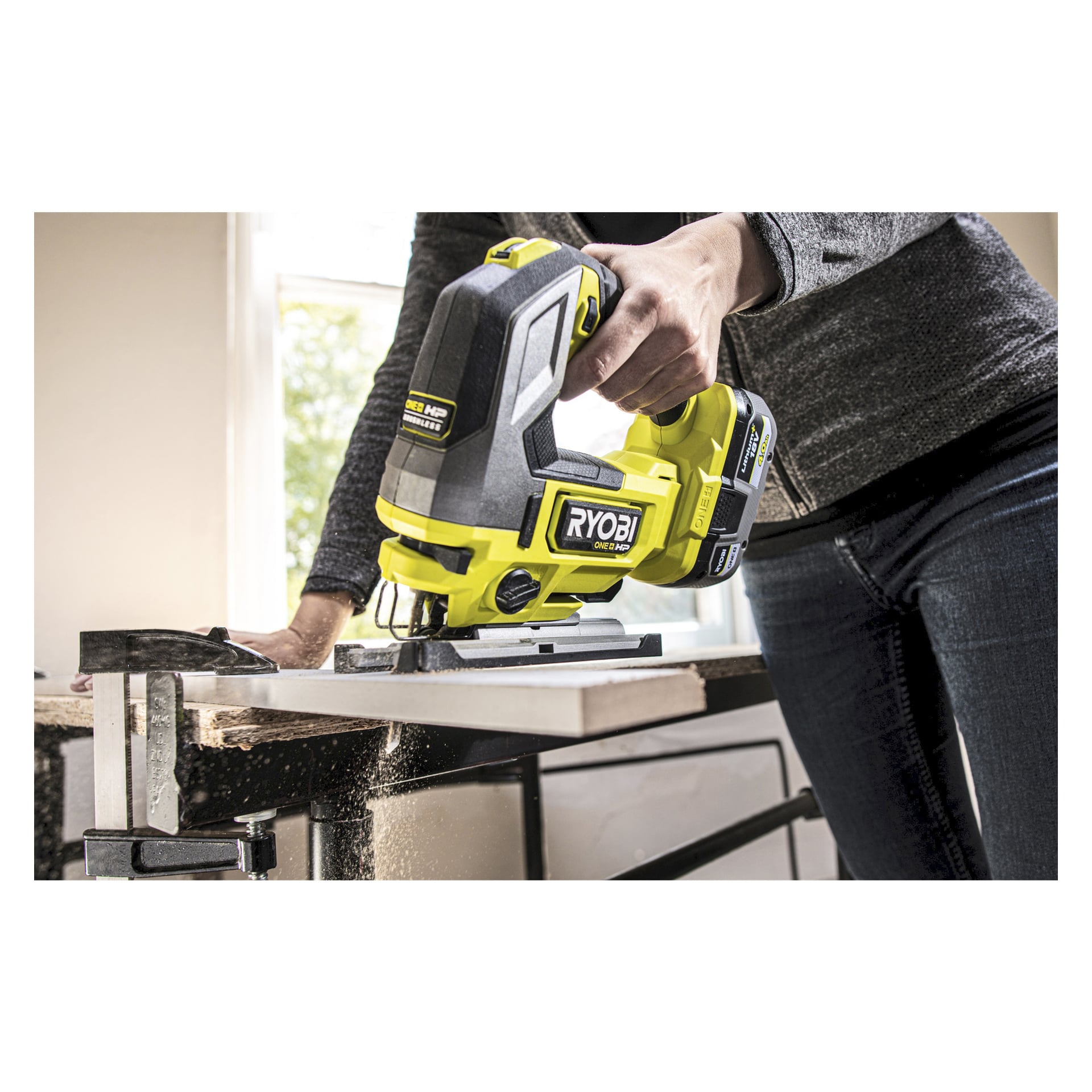 Osoba obsługuje wyrzutnię Ryobi One+ HP RJS18X-0, tnąc kawałek drewna przymocowany do stołu warsztatowego. Trociny są rozrzucone wokół, a światło słoneczne wpada przez pobliskie okno.