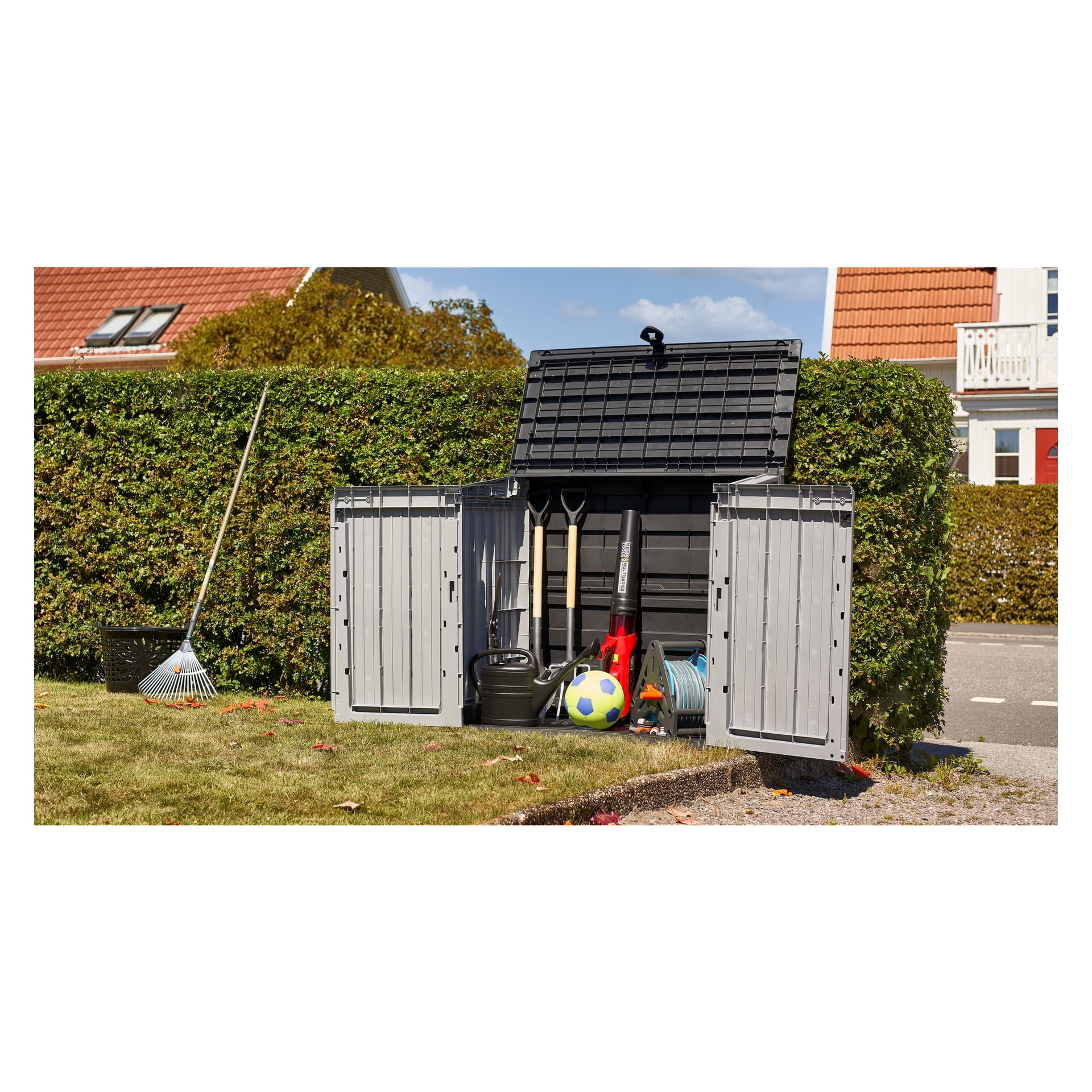 Den åpne Hard Head Storage har hageredskaper, en vannkanne, slange, fotball og diverse leker inni. En rake hviler inntil siden av enheten, som står på en gressplen avgrenset av en hekk med bolighus i bakgrunnen.