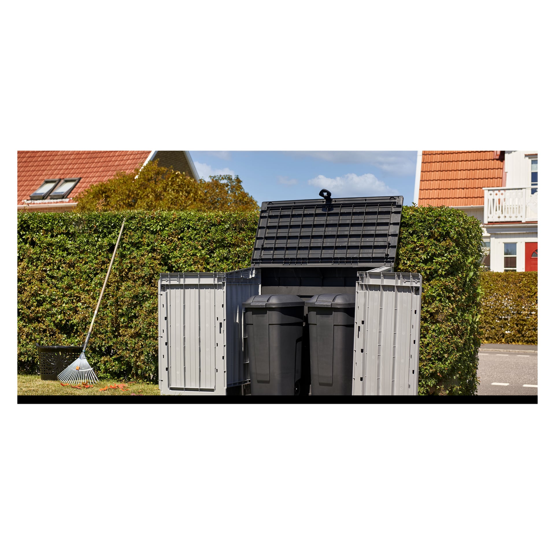 Et åpent Hard Head Storage rommer to svarte søppelkasser foran en grønn hekk. En rake og en svartløvpose ligger på gresset, og hus med røde tak sees i det fjerne.
