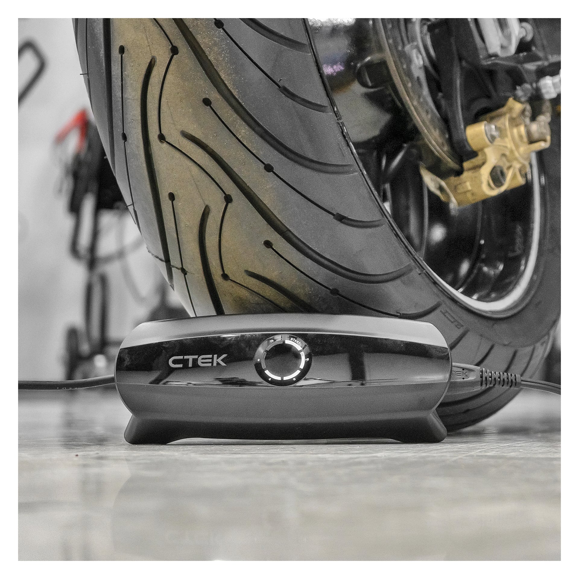 En detaljerad vy av ett motorcykeldäck på en blank yta, med CTEK-batteriladdaren CS ONE placerad direkt framför. De omgivande motorcykelkomponenterna och bakgrunden verkar mjukt ur fokus.