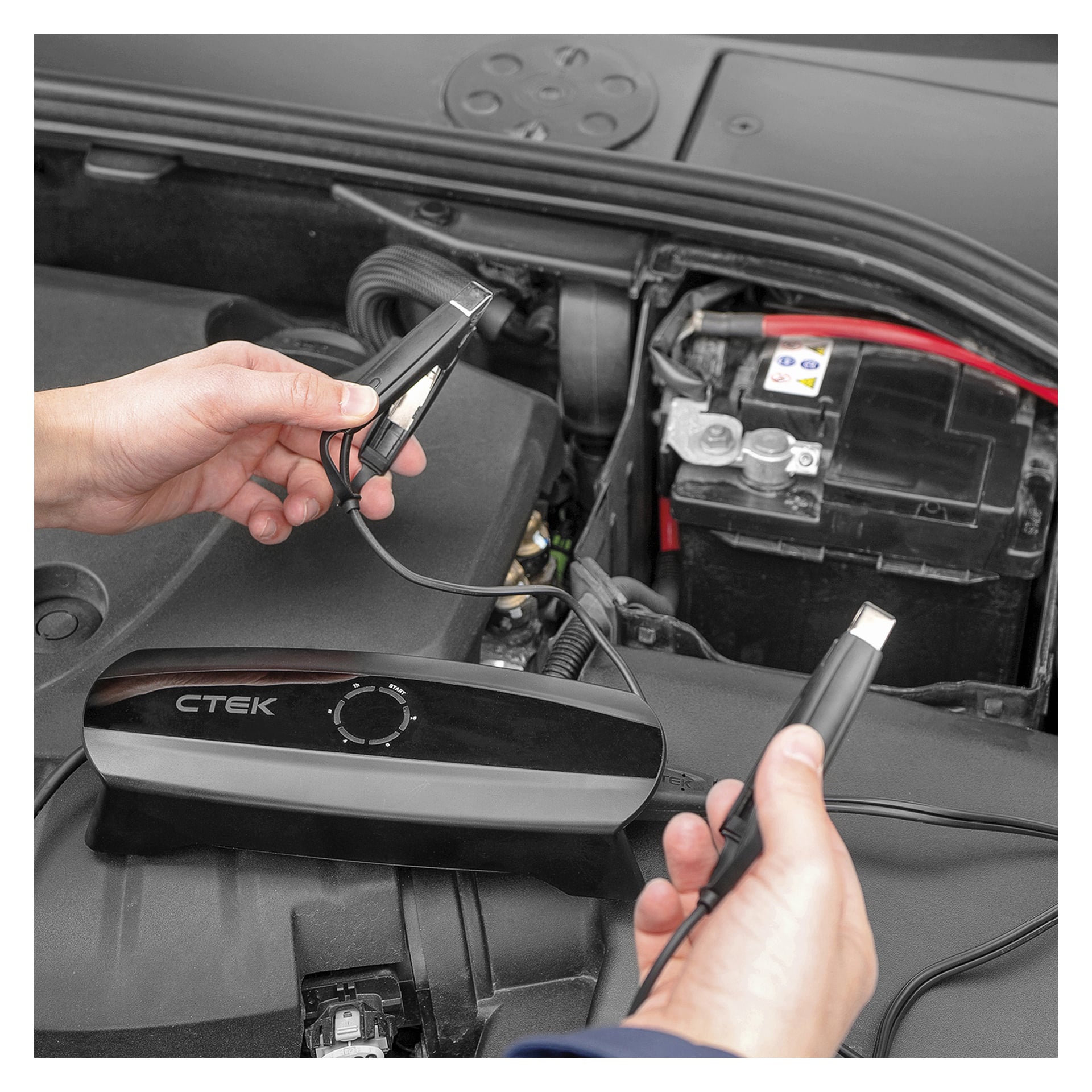 En person håller i batteriladdarklämmorna nära ett bilbatteri, medan en CTEK CS ONE batteriladdare vilar på motorn. Motorhuven är upphöjd, vilket exponerar motorkomponenter och anslutningskablar.