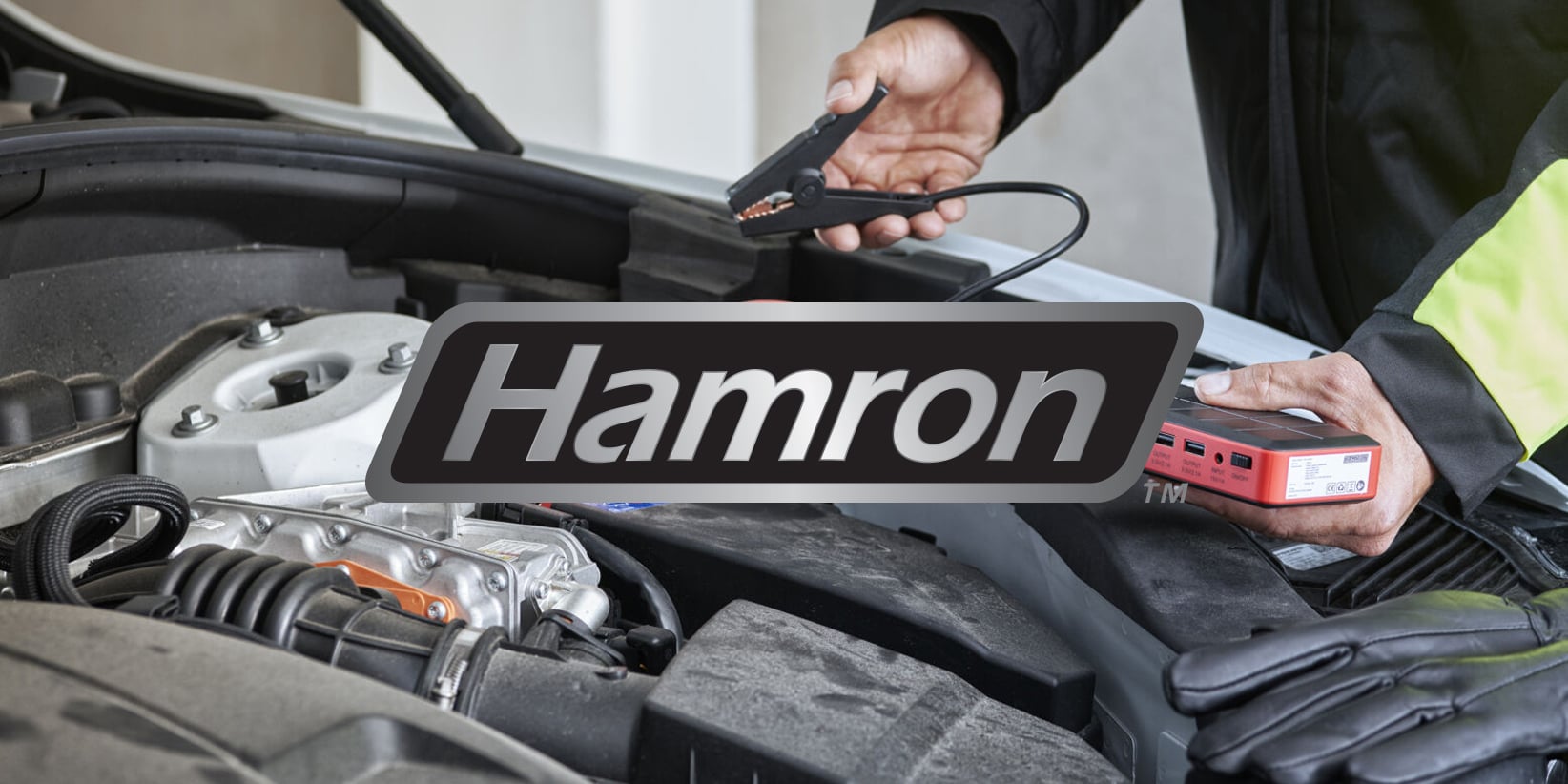 Någon håller startkablar och en batteritestare ovanför en bilmotor med ordet Hamron tydligt synligt över bilden.