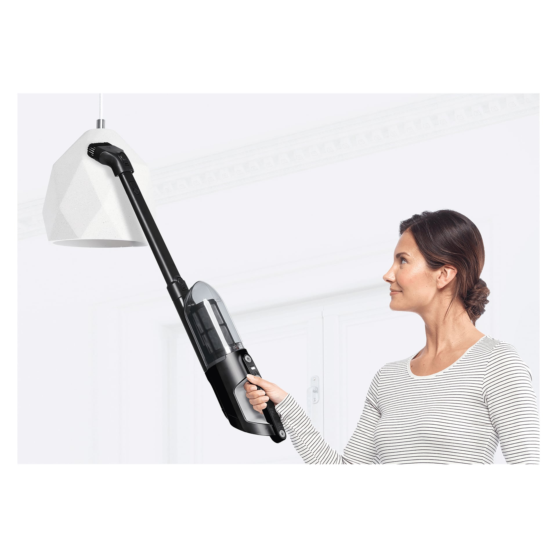 En kvinna som bär en randig långärmad skjorta använder en Bosch Upright dammsugare med förlängningsrör för att rengöra en vit geometrisk hänglampa.