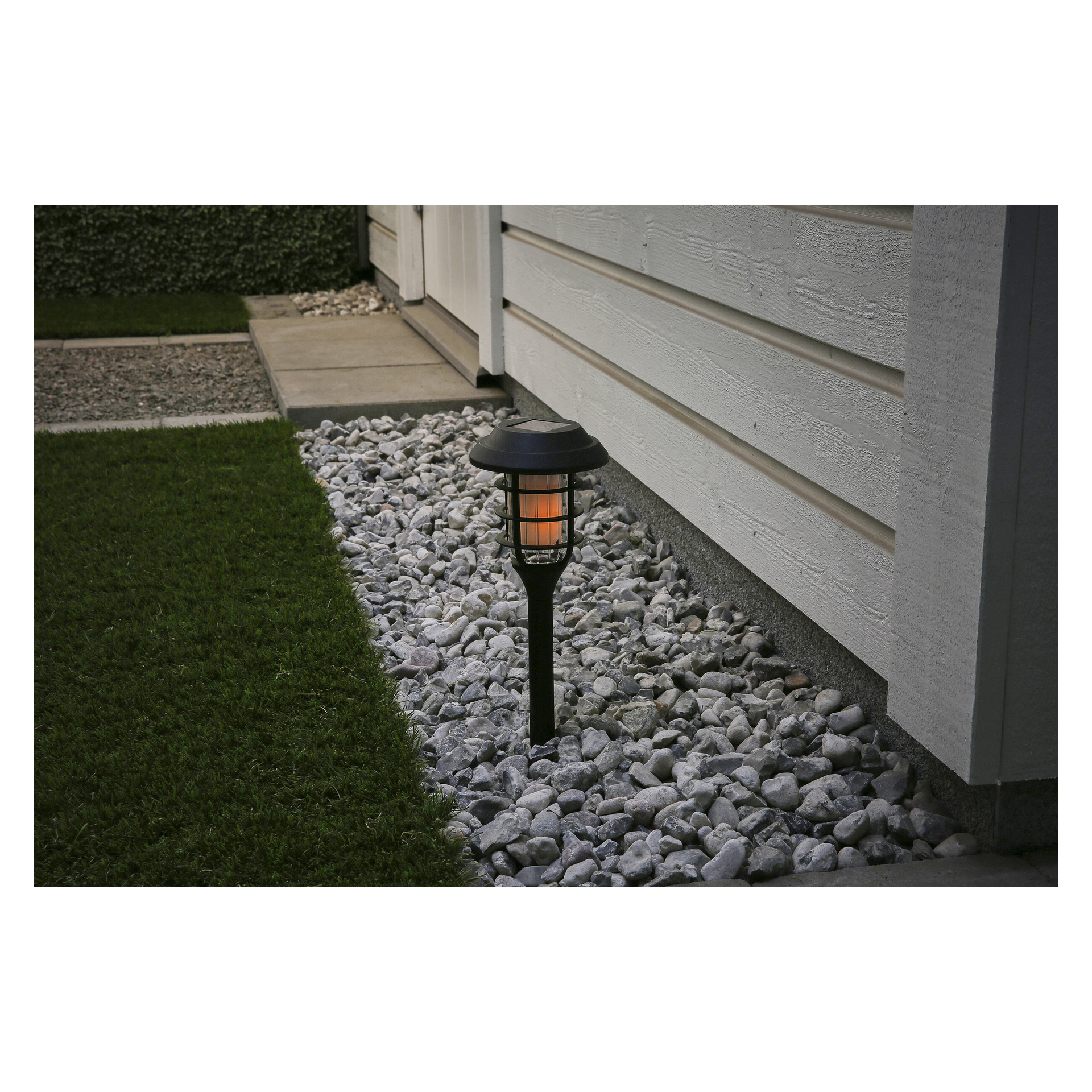 Lampa LED Anslut Solar Cell emitująca pomarańczowy blask jest umieszczona na szarych skałach obok białego budynku, otoczona zieloną trawą i wybrukowanym chodnikiem.