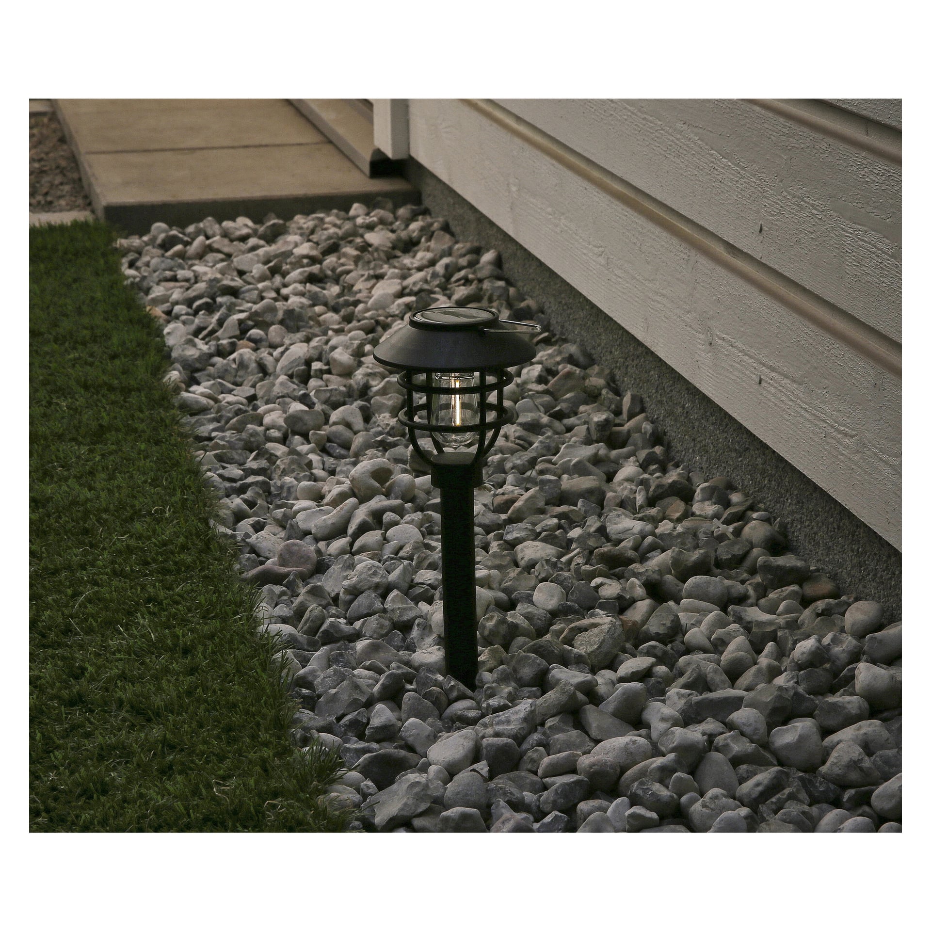 En Anslut solcellslampa med LED är placerad bland grå stenar bredvid husets yttervägg, med grönt gräs synligt på vänster sida av scenen.