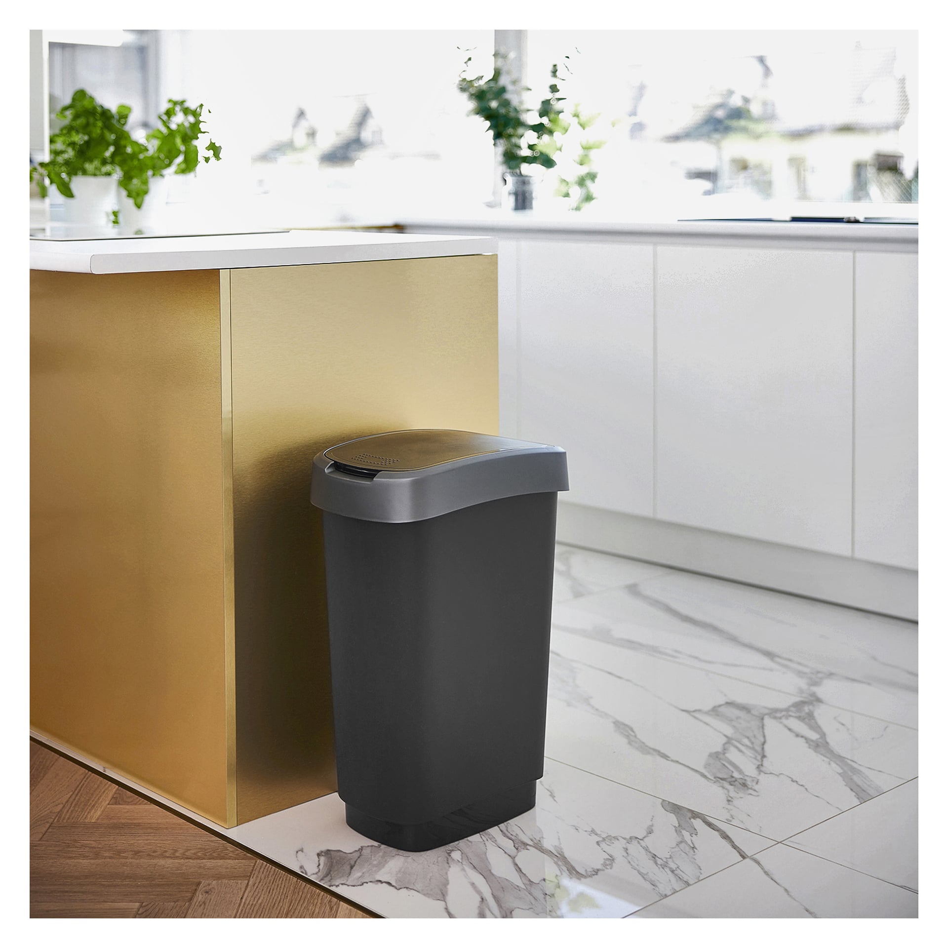 En Hard Head Waste-behållare med svart botten och grått lock står bredvid en guldfärgad köksbänk i ett elegant kök med vita skåp, gröna krukväxter och marmorgolv.