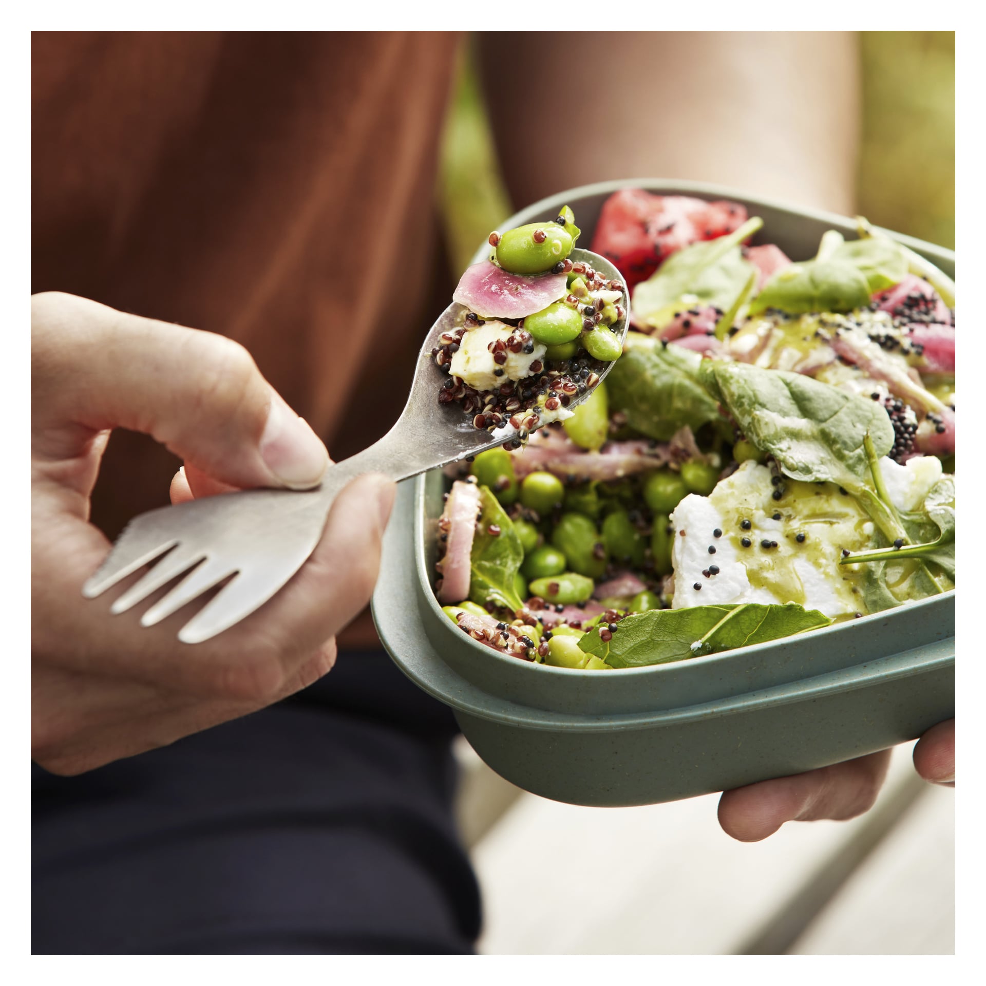 Någon äter en sallad gjord på quinoa, spenat, fetaost, ärtor och inlagd lök från en grön behållare med hjälp av en Light My Fire Spork. Scenen har en suddig bakgrund och en del av en brun skjorta syns.