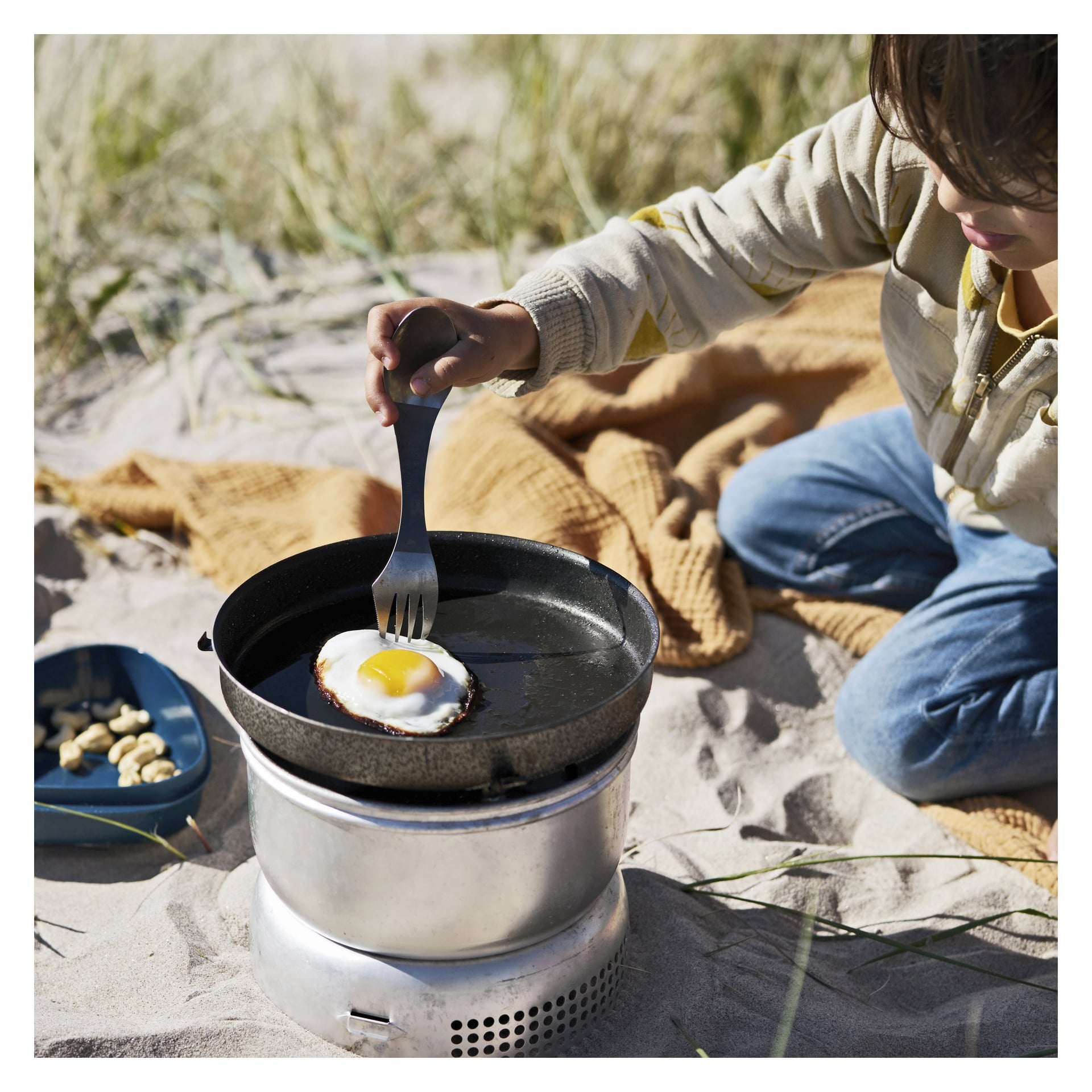 Ett barn förbereder ett ägg med solen uppåt i en svart panna med en Light My Fire Spork på en portabel spis på stranden, med en gul filt och en blå tallrik mat i närheten.