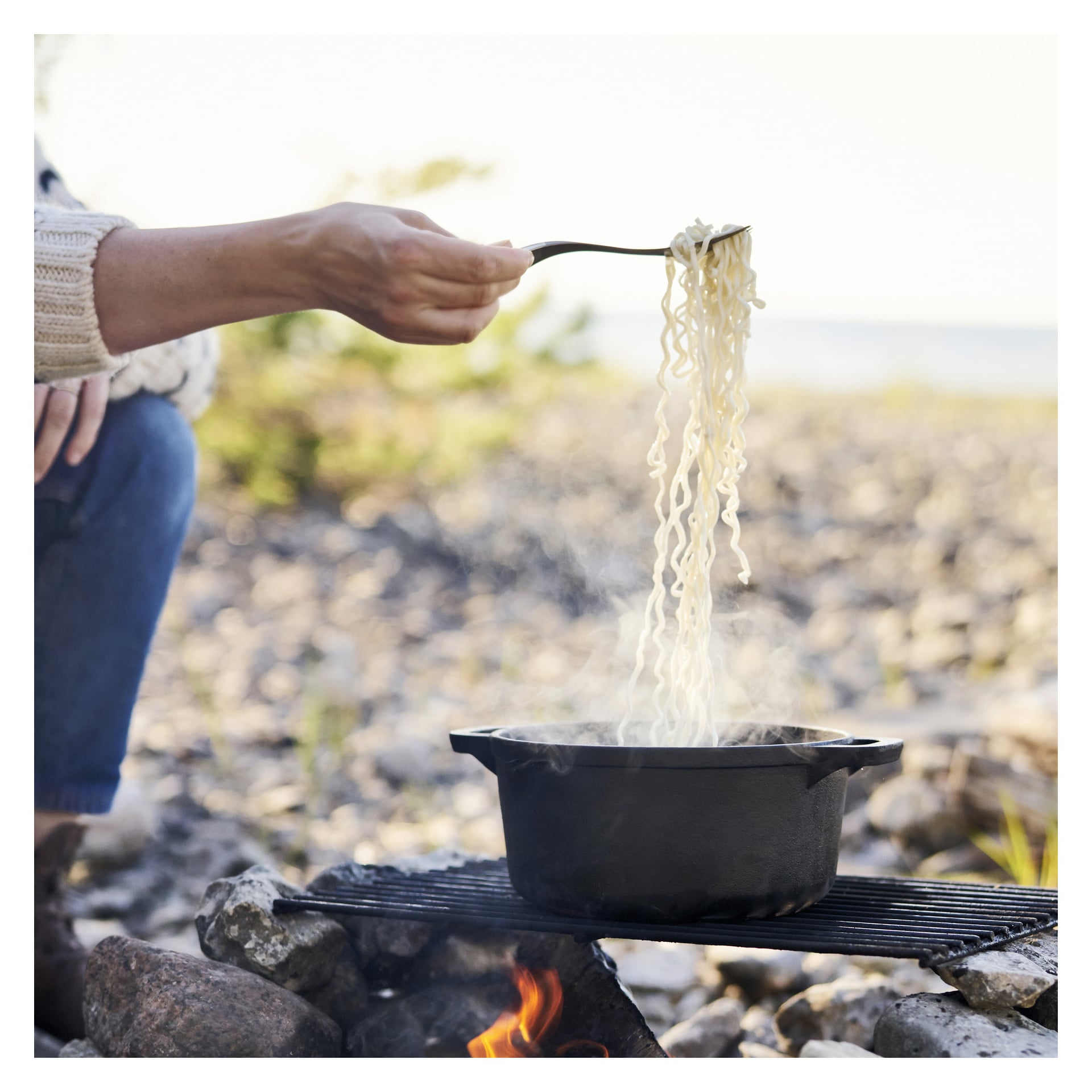 Någon använder en Light My Fire Spork för att skopa upp kokta nudlar från en ångande gryta, som står på en grill ovanför en lägereld, omgiven av stenar med en suddig naturlig bakgrund.