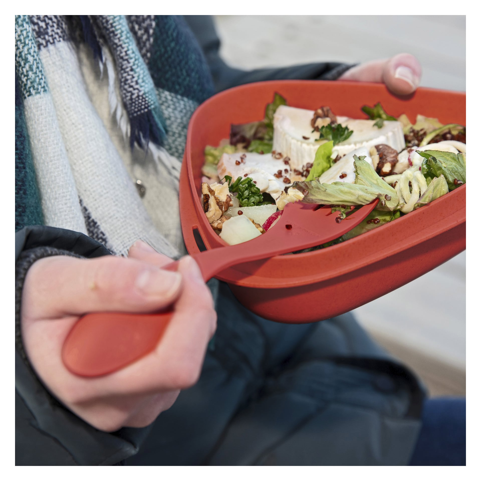 Noen kledd i grønn jakke og blått skjerf holder en rød salatbolle med grønnsaker, ost og nøtter, og spiser med en matchende rød Spork fra Light My Fire. Personens ansikt er ute av syne.