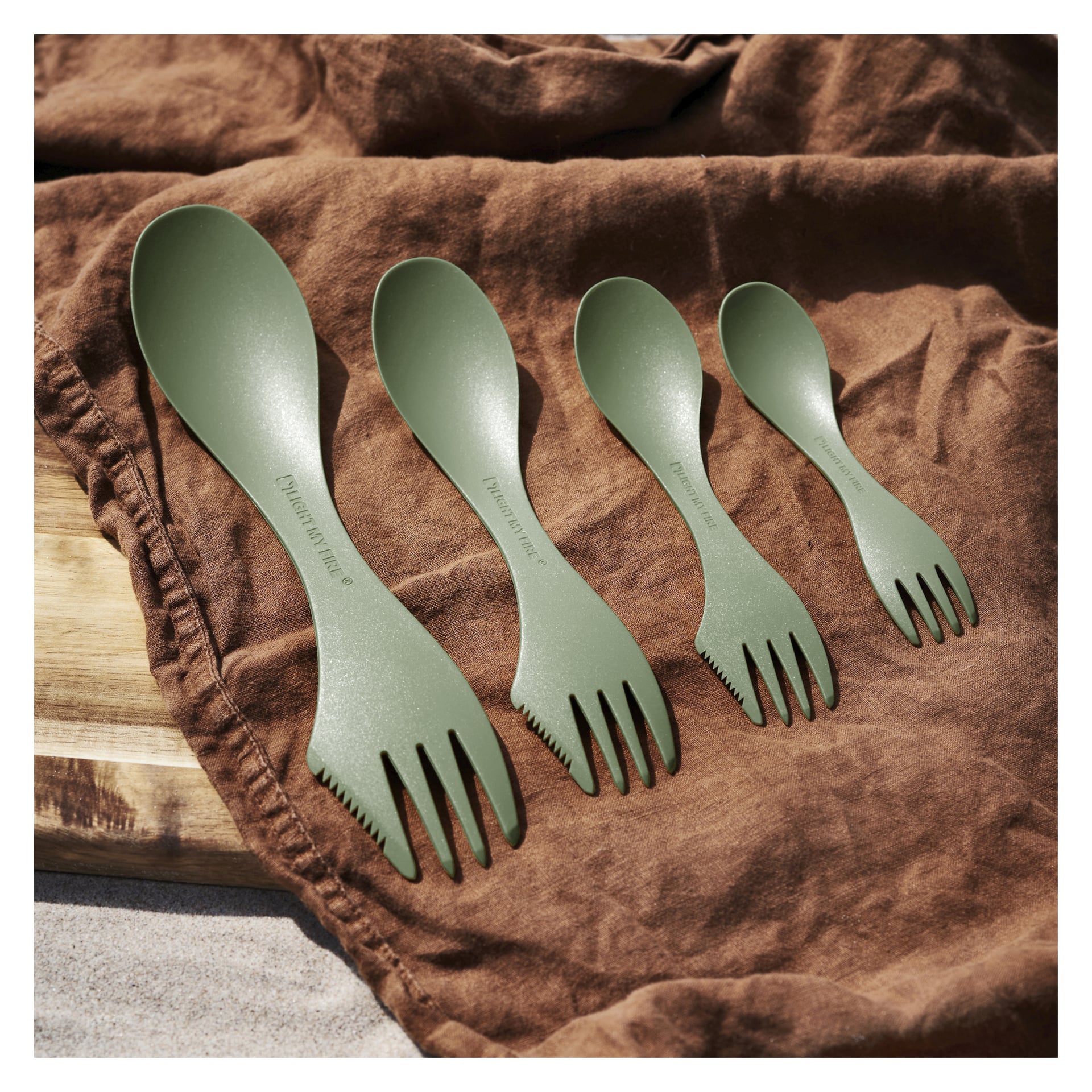 Et sett med fire grønne Light My Fire Spork-bestikk i forskjellige størrelser er stilt opp på et brunt stoff, med en treplate delvis synlig bak dem.