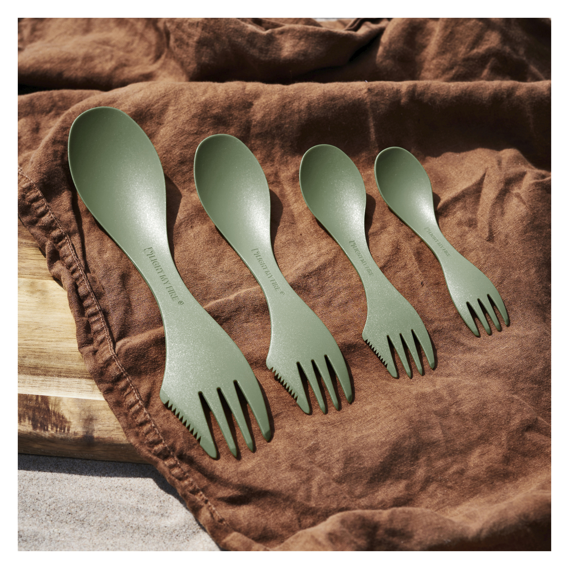 Neljä vihreää Light My Fire Spork -välinettä eri kokoisina on asetettu ruskealle kankaalle, ja niiden takana näkyy osittain puinen lauta.