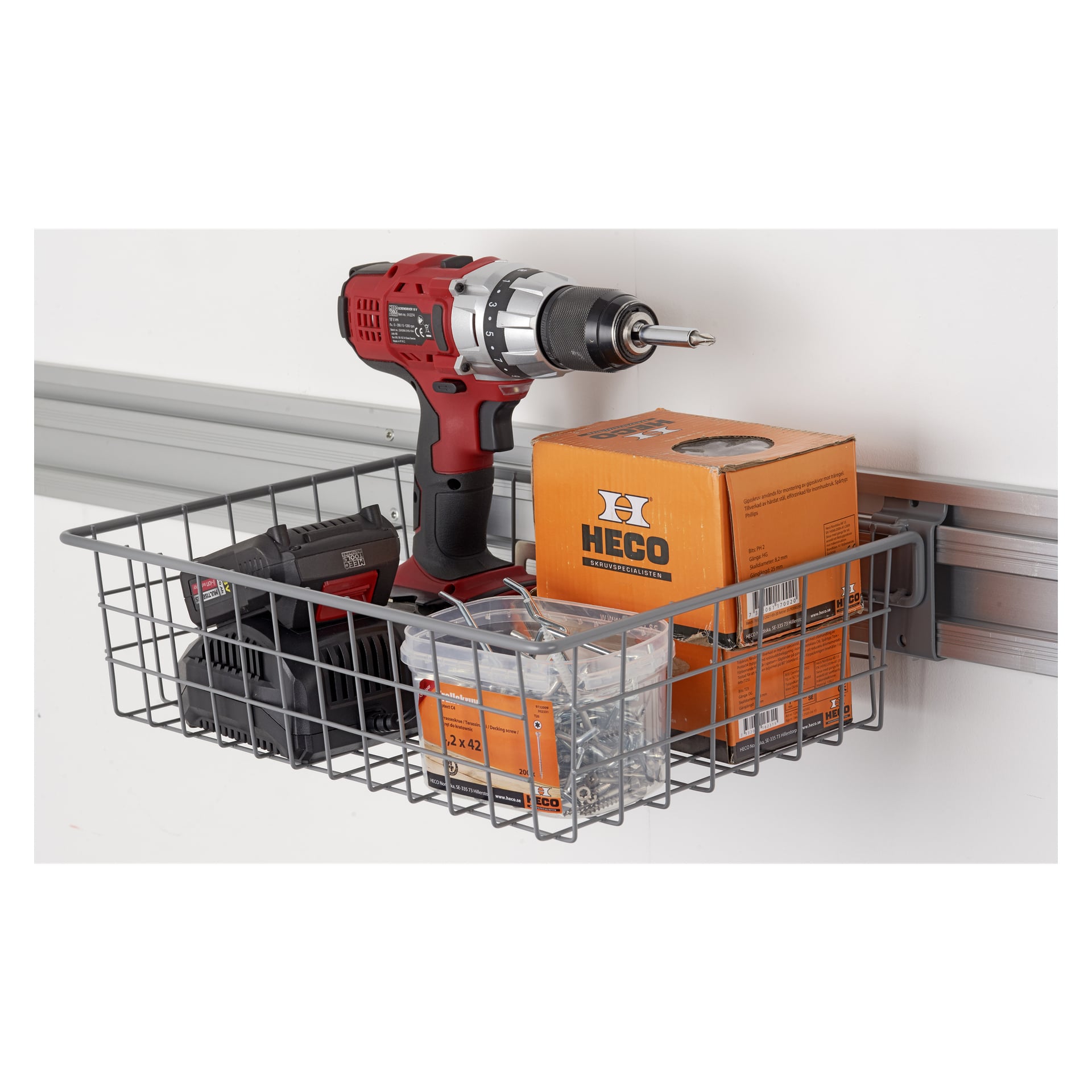 En metallkorg för hård huvudtråd är fäst vid en väggskena och förvarar prydligt en röd elektrisk borrmaskin, dess batteri, en laddare, en orange HECO-låda och en behållare fylld med skruvar eller fästen. Arrangemanget är ordnat och välorganiserat.