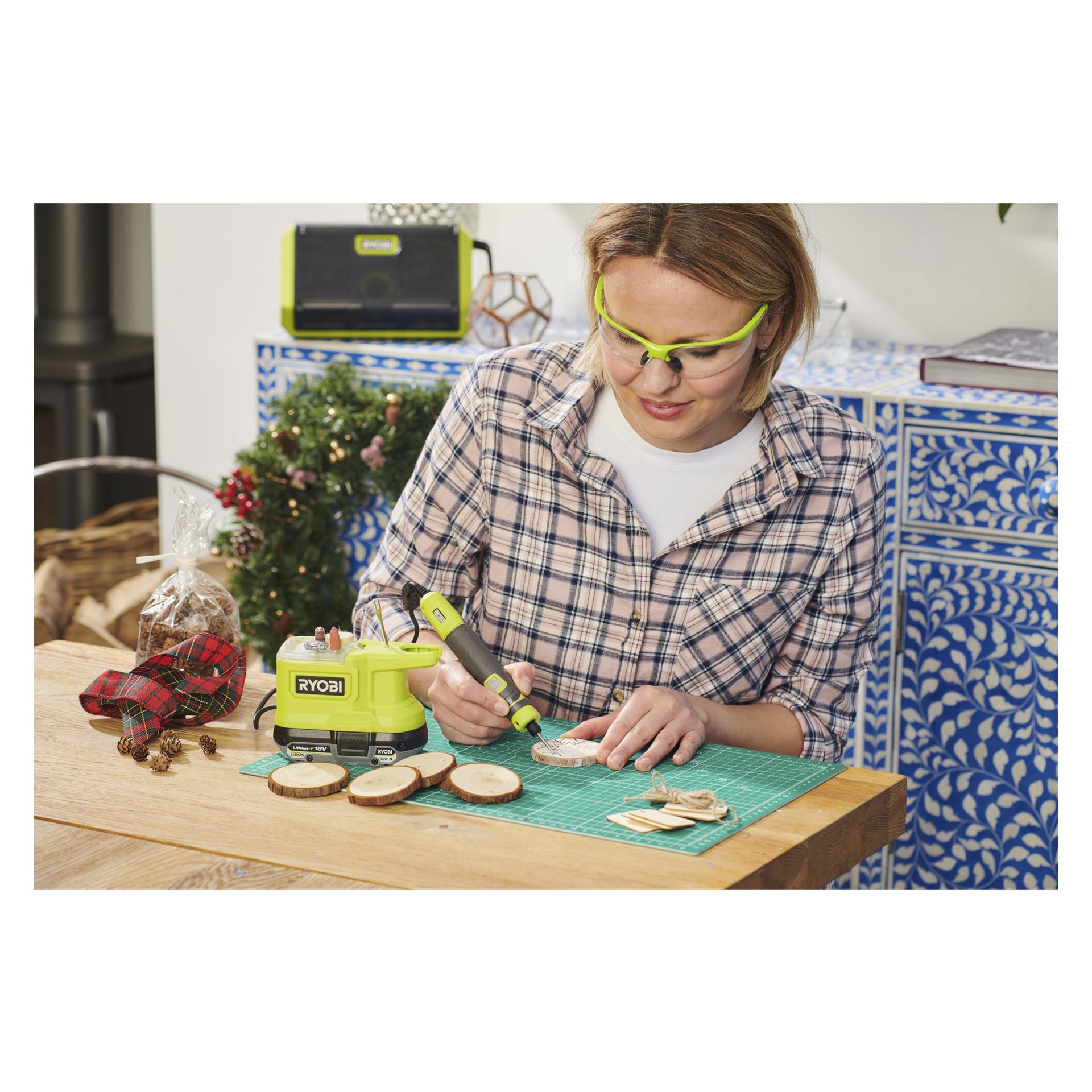 En person med skyddsglasögon graverar träskivor på ett bord med hjälp av Ryobi One+ Multi-tool Mini RRT18-0. I bakgrunden finns juldekorationer, en inslagen present och blå skåp med mönster.