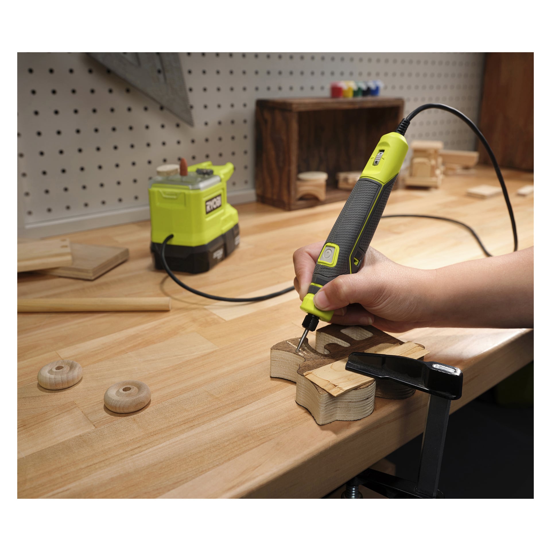 En hand manövrerar den gröna och svarta Ryobi One+ Multi-tool Mini RRT18-0 och graverar en design i en träbit som hålls på en arbetsbänk. Träytan är utspridd med olika verktyg och träföremål i en verkstadsmiljö.