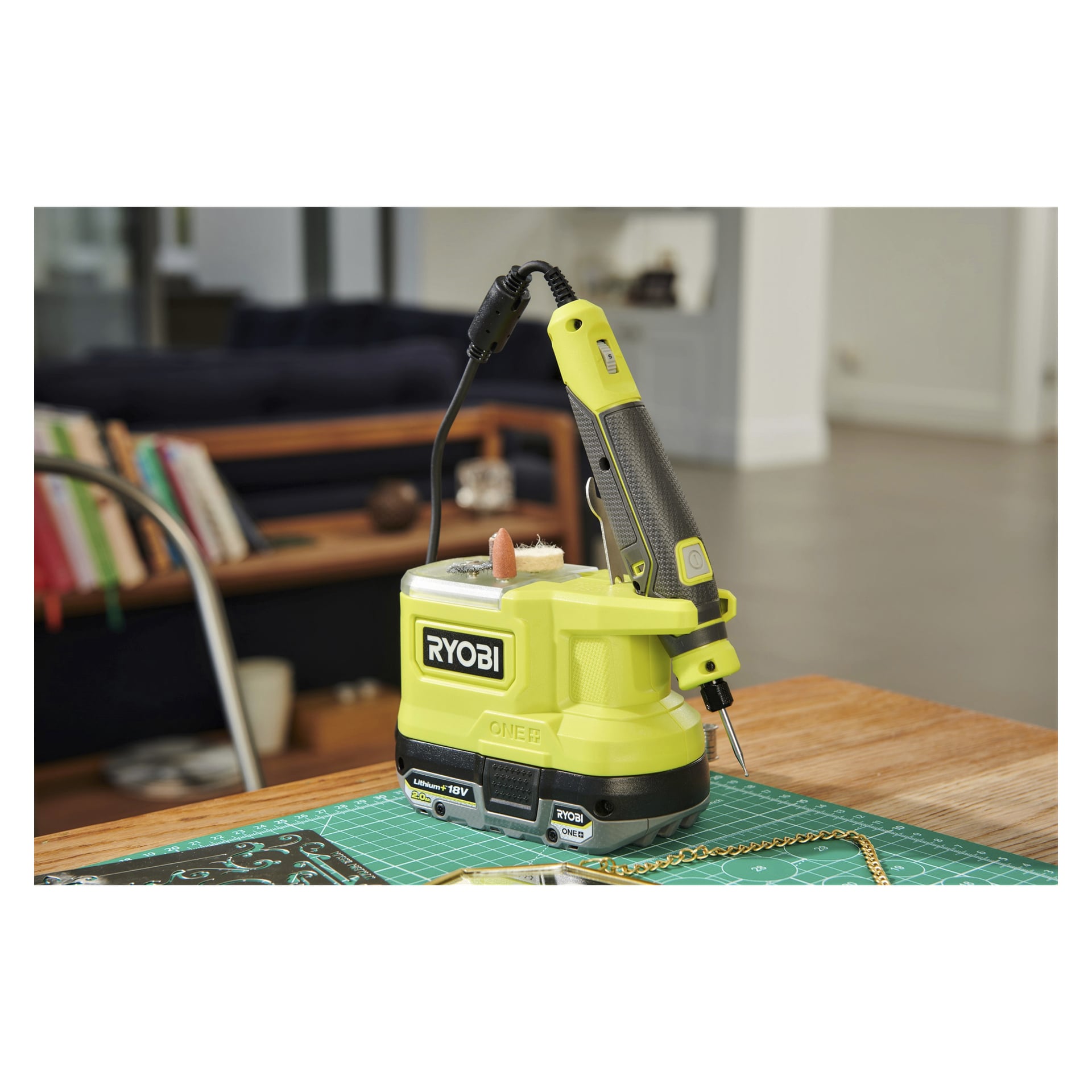 En Ryobi One+ Multitool Mini RRT18-0 står på ett träbord, fäst vid sin kraftbas med olika hantverksmaterial och en skärmatta runt omkring. Bakgrunden avslöjar en del av ett vardagsrum.