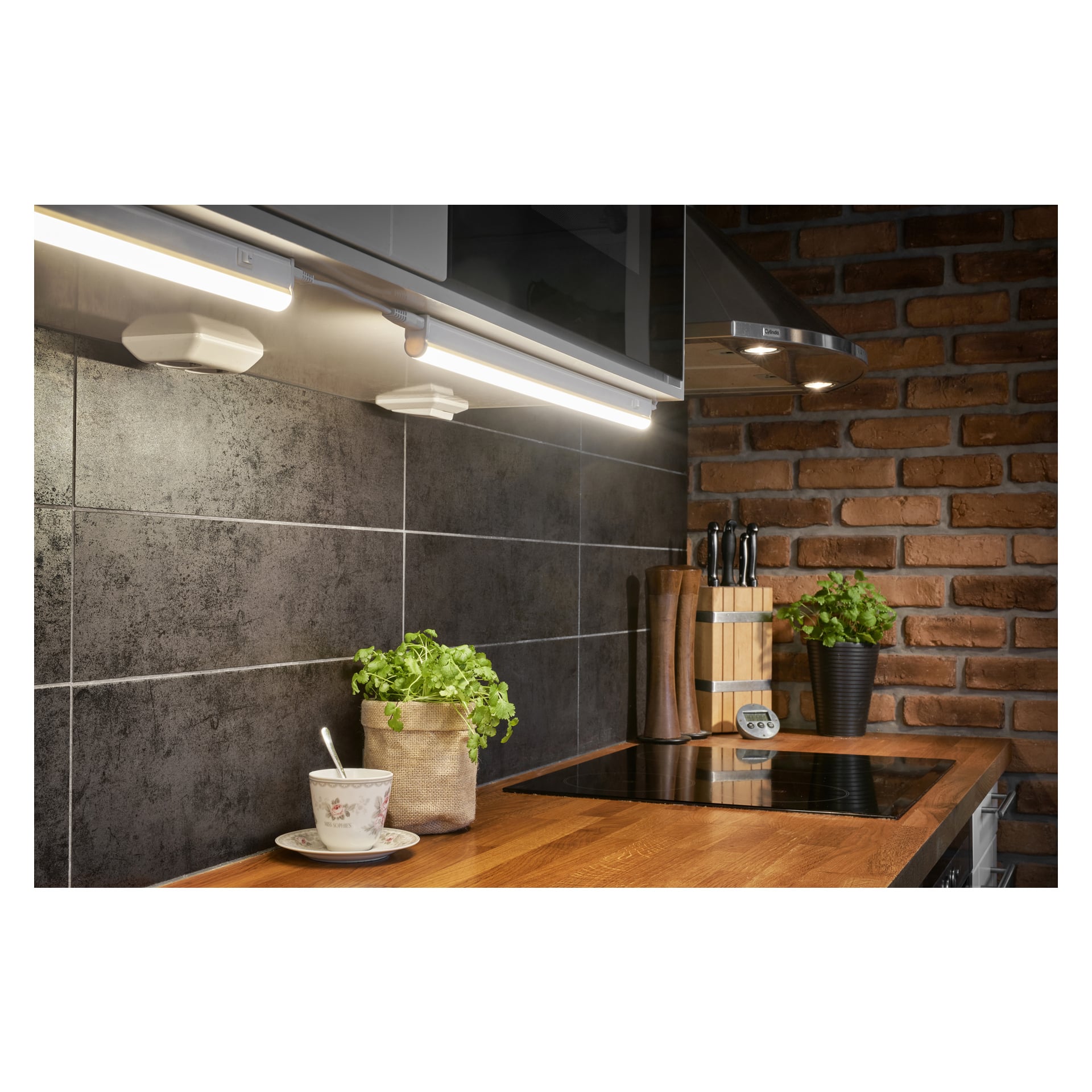 En modern köksbänk med kopp och fat, krukväxter, knivar i en träkloss, elektrisk spis och Anslut Worktop Light LED installerad under skåpen. Stänkskyddet har mörka kakel och det finns en synlig tegelvägg.